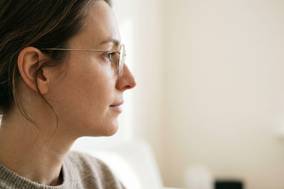 Side profile of an adult wearing thin wire-rimmed glasses and a knit sweater, gazing ahead with a calm expression in a softly lit room.